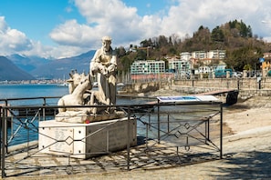 Une statue représentant un homme et un cheval, placée sur un piédestal au bord de l’eau, avec des bâtiments et des montagnes en arrière-plan.