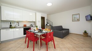 A modern kitchen with white cabinets, a dining table with red chairs, and a grey sofa in the living area. - Le Betulle Garden in Laveno (Laveno Mombello)