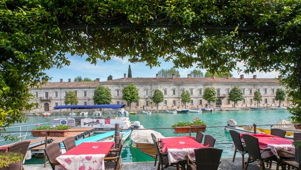Une terrasse au bord de la rivière, avec des tables et des chaises, offrant une vue sur un bâtiment historique situé de l’autre côté de l’eau, et des bateaux amarrés à proximité.