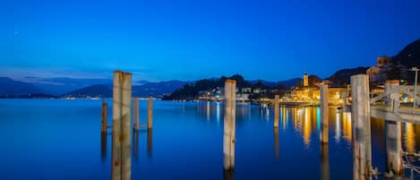 A calm waterfront with wooden posts and a bridge, reflecting city lights and buildings in the background.