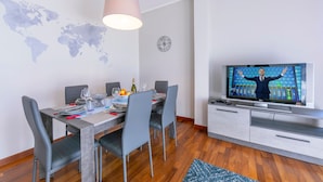 Un coin repas avec une table en bois, des chaises grises et une carte du monde accrochée au mur. Un téléviseur est fixé au mur et affiche l’image d’une personne.