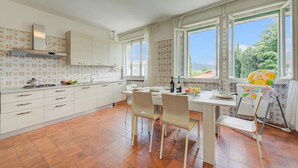 A modern kitchen with white cabinets, a dining table with chairs, and large windows offering a view of greenery. - Margherita Near Lake-child Friendly (Toscolano Maderno)
