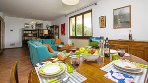 Un comedor con una mesa de madera lista para una comida, un sofá al fondo y vistas al exterior a través de amplios ventanales.