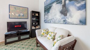 Een woonkamer met een televisie waarop 'NETFLIX' staat, een boekenkast, een witte bank met bloemenkussens, en een groot schilderij van een vuurtoren.