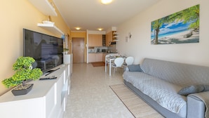 A modern living room with a flat-screen TV, a grey sofa, a dining table with chairs, and a kitchen in the background. - Kristall Lago Pool View (Desenzano del Garda)