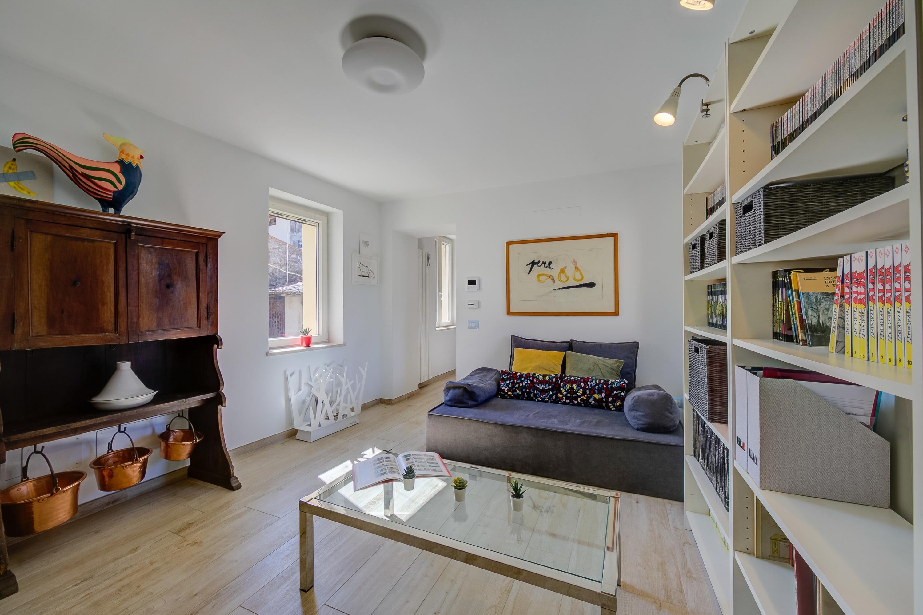 A cozy living room with a wooden cabinet, a glass coffee table, a sofa with cushions, and a bookshelf filled with books and decorative items.