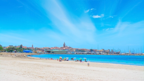 A beach with clear blue water, a sandy shore, and a cityscape in the background. - Bilocale Vista Mare Giorgio (Alghero)