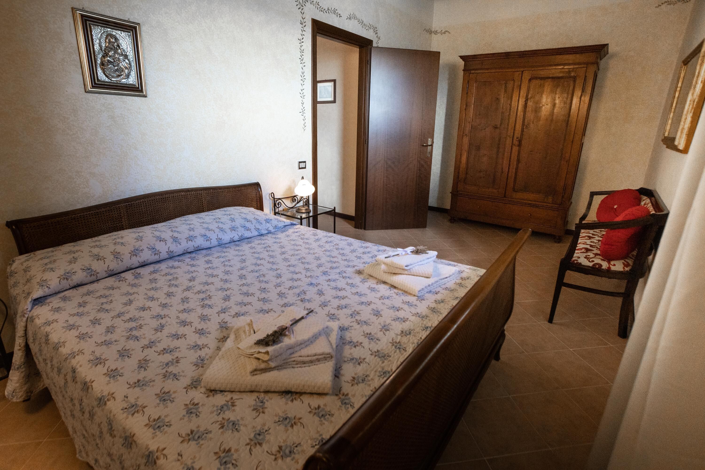 A bedroom with a bed, a wooden wardrobe, a chair, and a nightstand with a lamp.