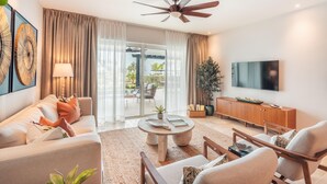 A modern living room with a ceiling fan, a flat-screen TV, a sofa, a coffee table, and a sliding glass door leading to an outdoor area. - Apartment w Pool Ocean View at Punta Palmera BA5 (Punta Cana)