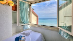 A hotel room with a large window offering a view of the sea, a bed with a white bedspread, a bedside table with a book and a glass of water, and a wall-mounted lamp. - Il Bijou Di Rossella - Beach Front (Alassio)