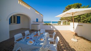 Un espace de restauration extérieur baigné de lumière, avec une table dressée pour quatre personnes, des chaises blanches et un grand parasol.