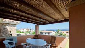 A covered outdoor patio with a round table and chairs, stone wall, and a view of buildings and mountains in the background. - Le Dimore di Nettuno 4 With Pool (Olbia)