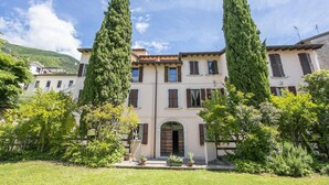 A large, multi-story building with a symmetrical facade, featuring a central entrance with an archway, flanked by tall, slender trees. - Berny 150m From Lake (Salò)