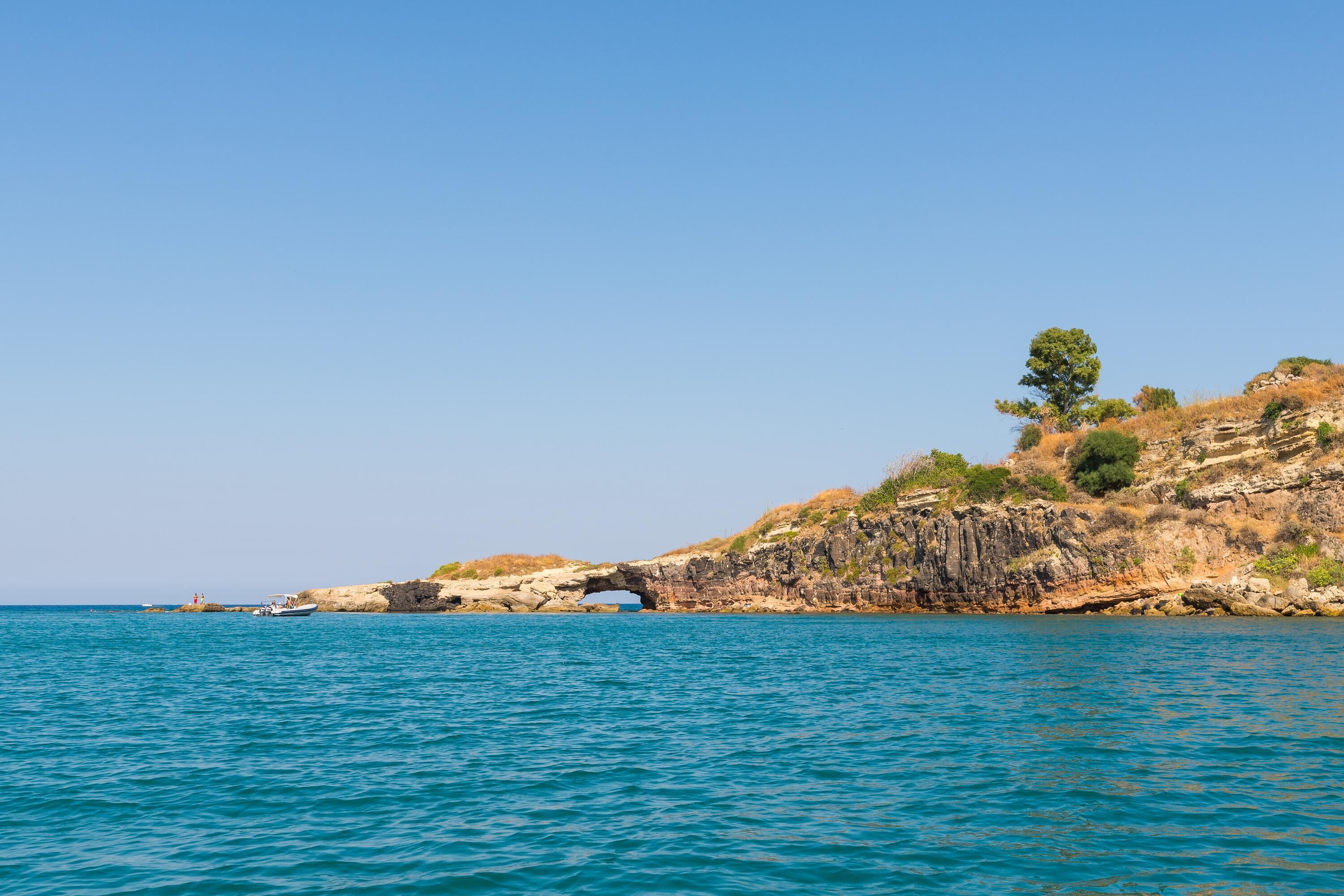 A coastal scene with a clear blue sky, a rocky cliff, and a boat in the water.
