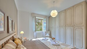 Ein Schlafzimmer mit Bett, Nachttisch, Lampe, Kleiderschrank und einem Fenster mit Blick ins Grüne.