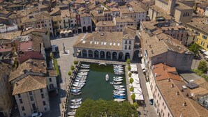 An aerial view of a historic town with a central square, boats docked in a marina, and traditional buildings with arched windows.