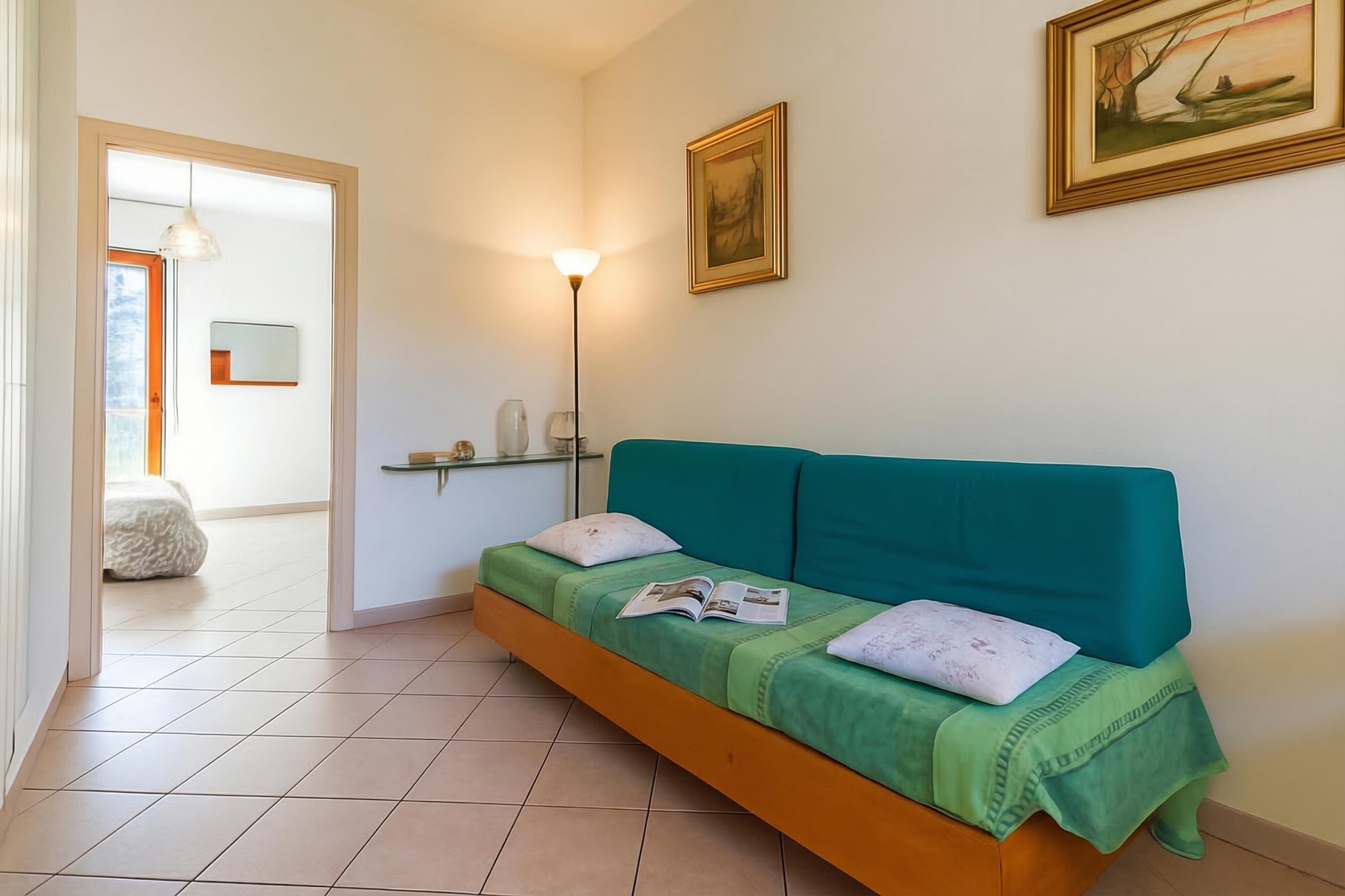 A room with a green sofa, a lamp, a framed picture, and a bed in the background.