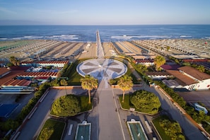 An aerial view of a coastal resort with a central circular plaza, multiple beachside villas, and a long pier extending into the ocean.
