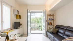 Un salon moderne avec un canapé en cuir noir, une table à manger avec des chaises, un téléviseur et un balcon offrant une vue sur la verdure.