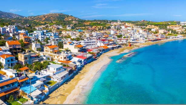 Un pueblo costero con edificios a lo largo de la playa, aguas azules cristalinas y un paisaje montañoso de fondo.