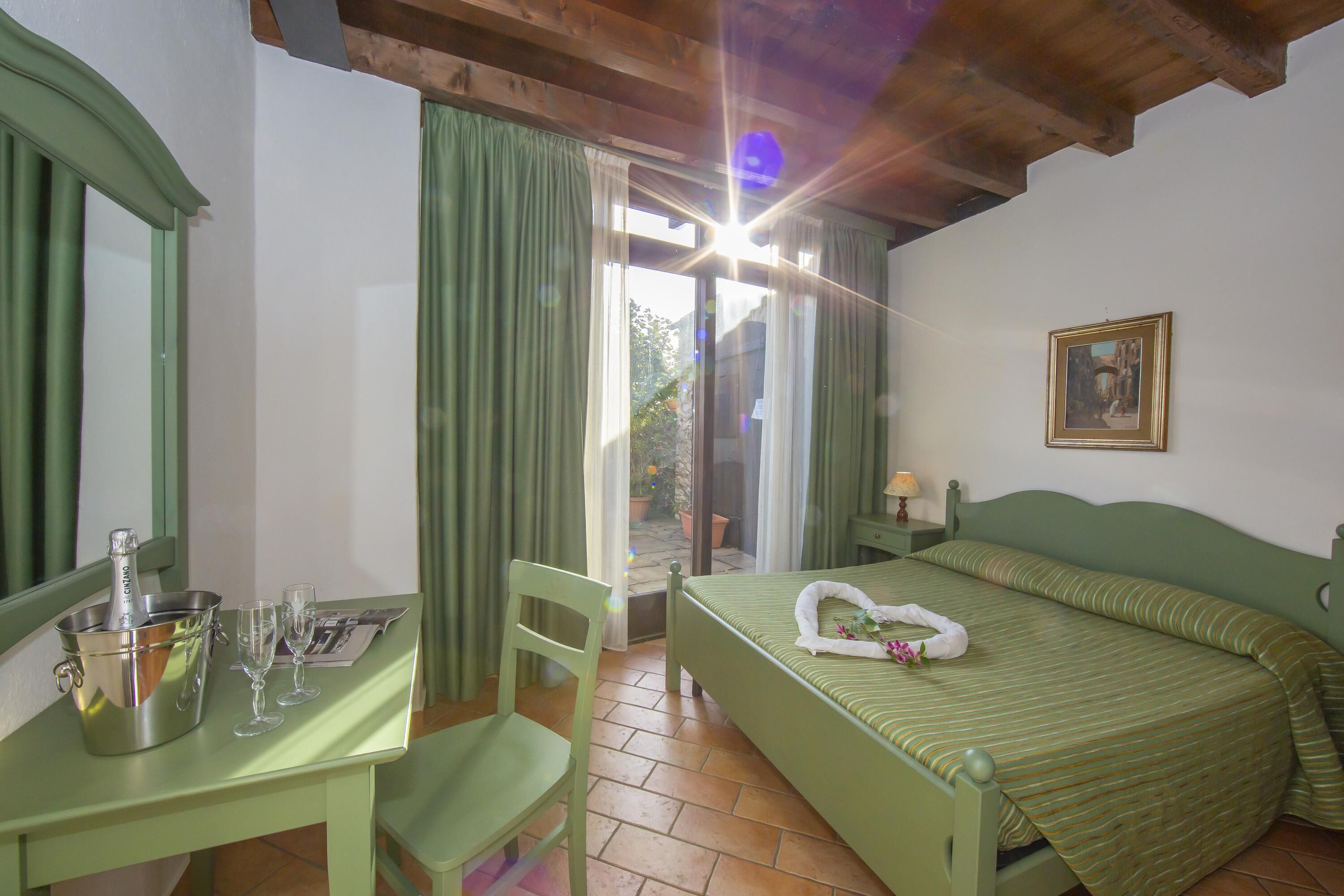 A room with a bed, a table with a bottle and glasses, a chair, and a painting on the wall.