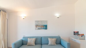 A light blue sofa with two pillows, a painting of boats on the wall, and a small cabinet with books. - Feel So Good in Bunthe (Olbia)