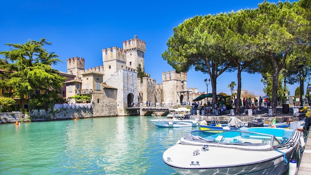 Un castillo histórico con un foso, barcos amarrados en el muelle y un cielo azul despejado.