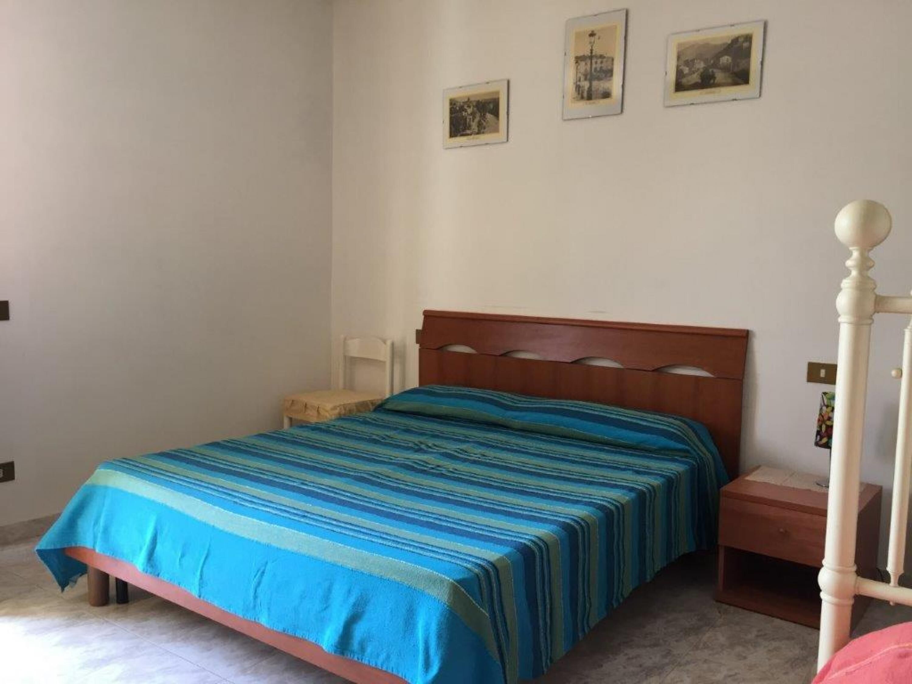 Ένα υπνοδωμάτιο με ένα κρεβάτι, ένα κομοδίνο, μια καρέκλα και τρεις φωτογραφίες σε κορνίζες στον τοίχο.