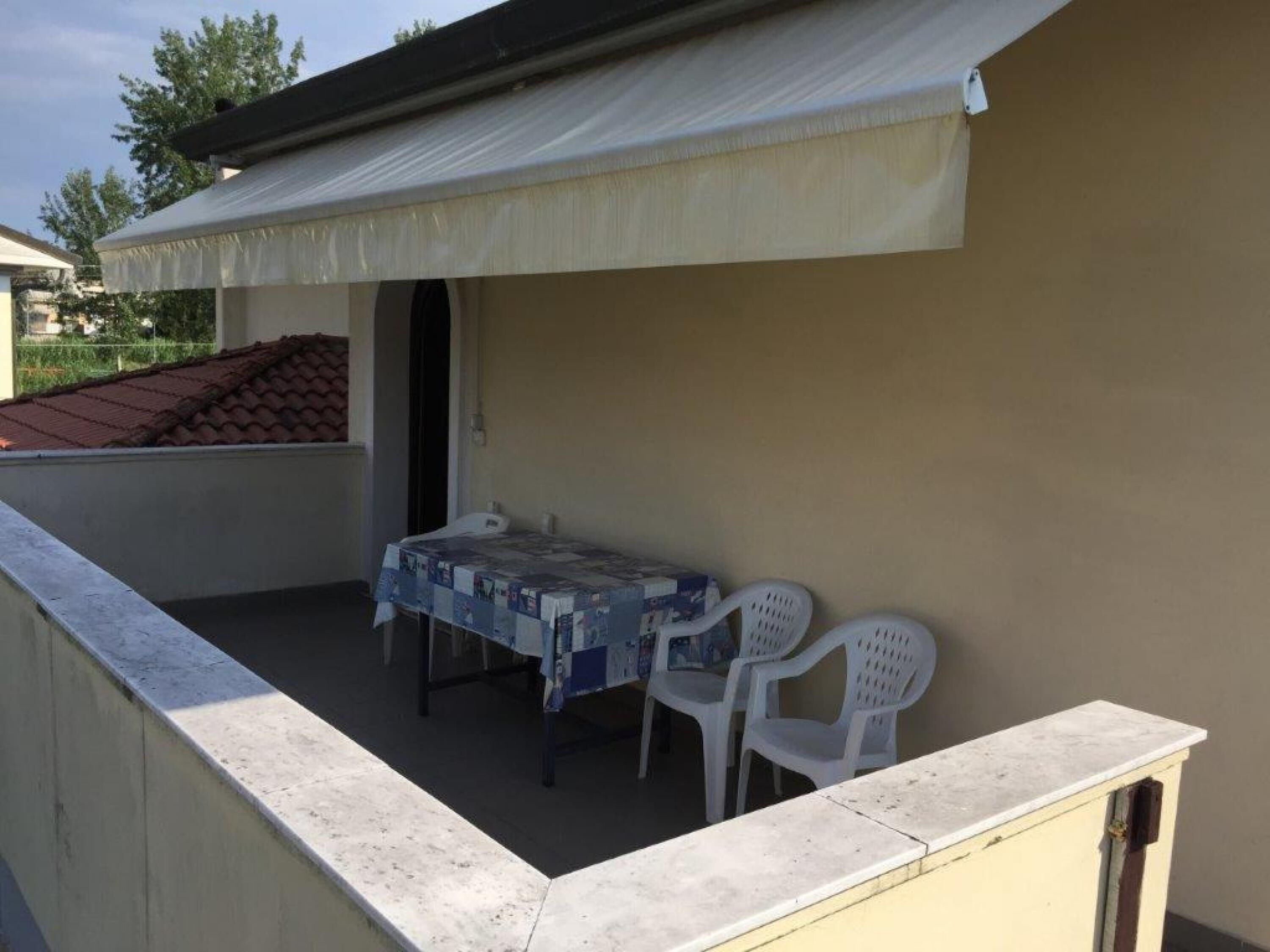A small outdoor patio with a table and two chairs, a covered area, and a view of a red-roofed building in the background.