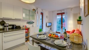 Une cuisine moderne avec des meubles blancs, une table à manger dressée avec des assiettes, des verres et un panier de fruits. Il y a un téléviseur accroché au mur et une fenêtre avec des rideaux.