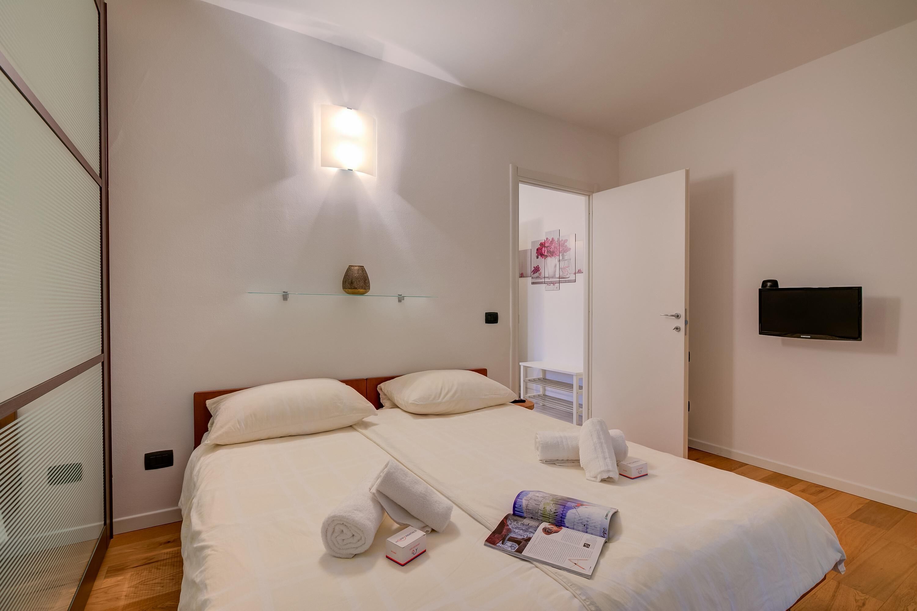 Una camera d'albergo con un letto, un comodino con un vaso, una TV a muro e una porta che conduce al bagno.