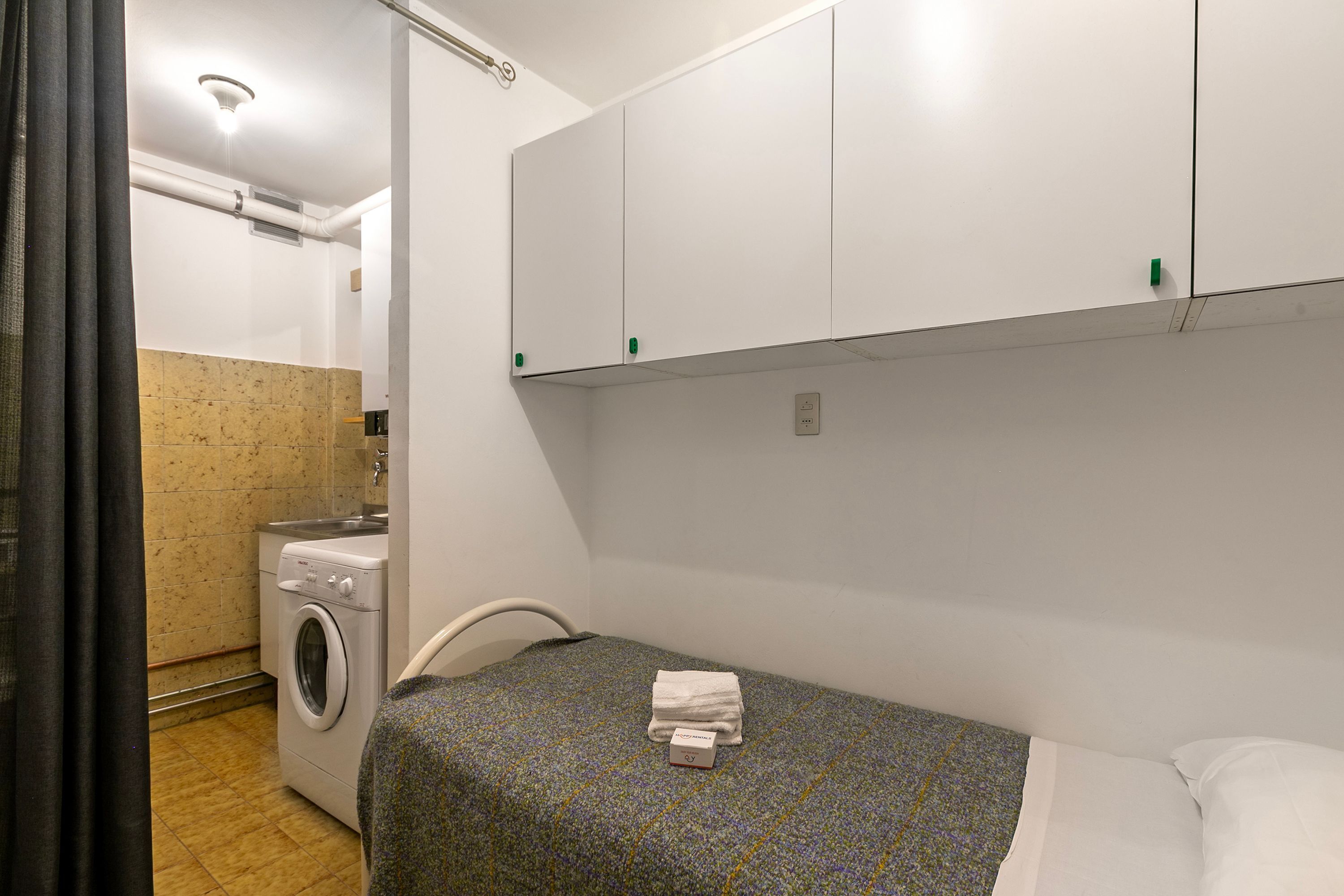 Une petite pièce propre, équipée d’un lit, d’une machine à laver et d’un coin cuisine.