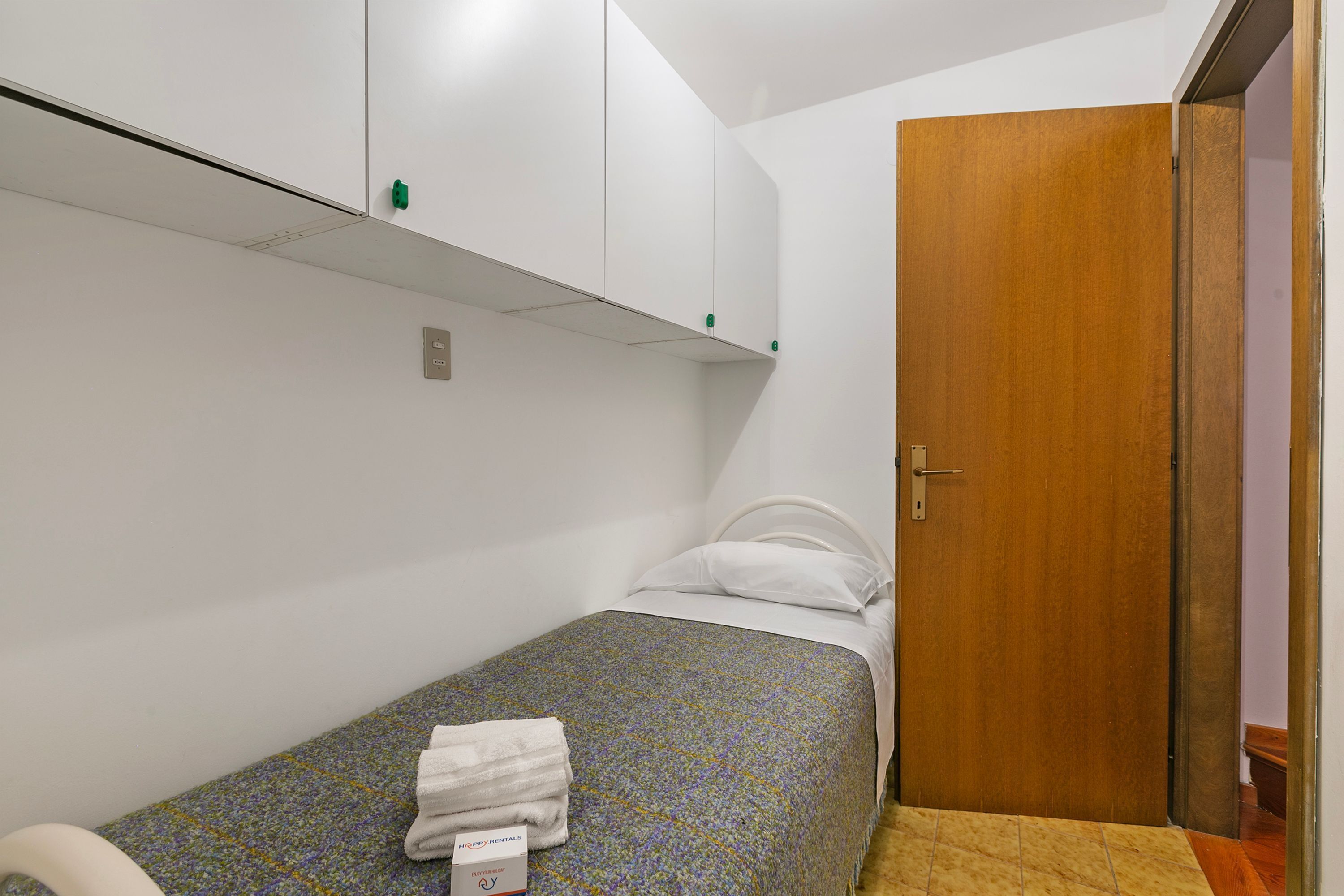 A small, single-bed room with a wooden door, a white wall, and a patterned bedspread.