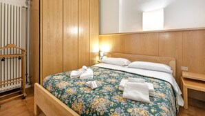 A hotel room with a wooden bed, a radiator, a wooden wardrobe, and a bedside table with a lamp. - Ca Cerere in Riva del Garda (Riva del Garda)