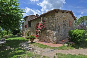 Un edificio in pietra con una porta rossa e finestre, circondato da un giardino con piante in vaso e un vialetto.