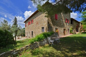 Un edificio in pietra con persiane rosse, un muro in pietra e un prato con una sedia in metallo.