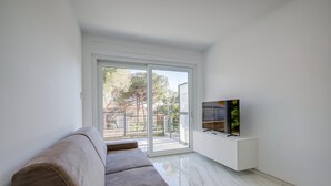 Una sala de estar moderna con un sofá, un televisor sobre un mueble blanco y un ventanal corredero que da a un balcón.