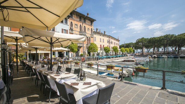 Un restaurant situé au bord de l’eau, avec une terrasse, des tables et des parasols, donnant sur un canal où naviguent des bateaux.