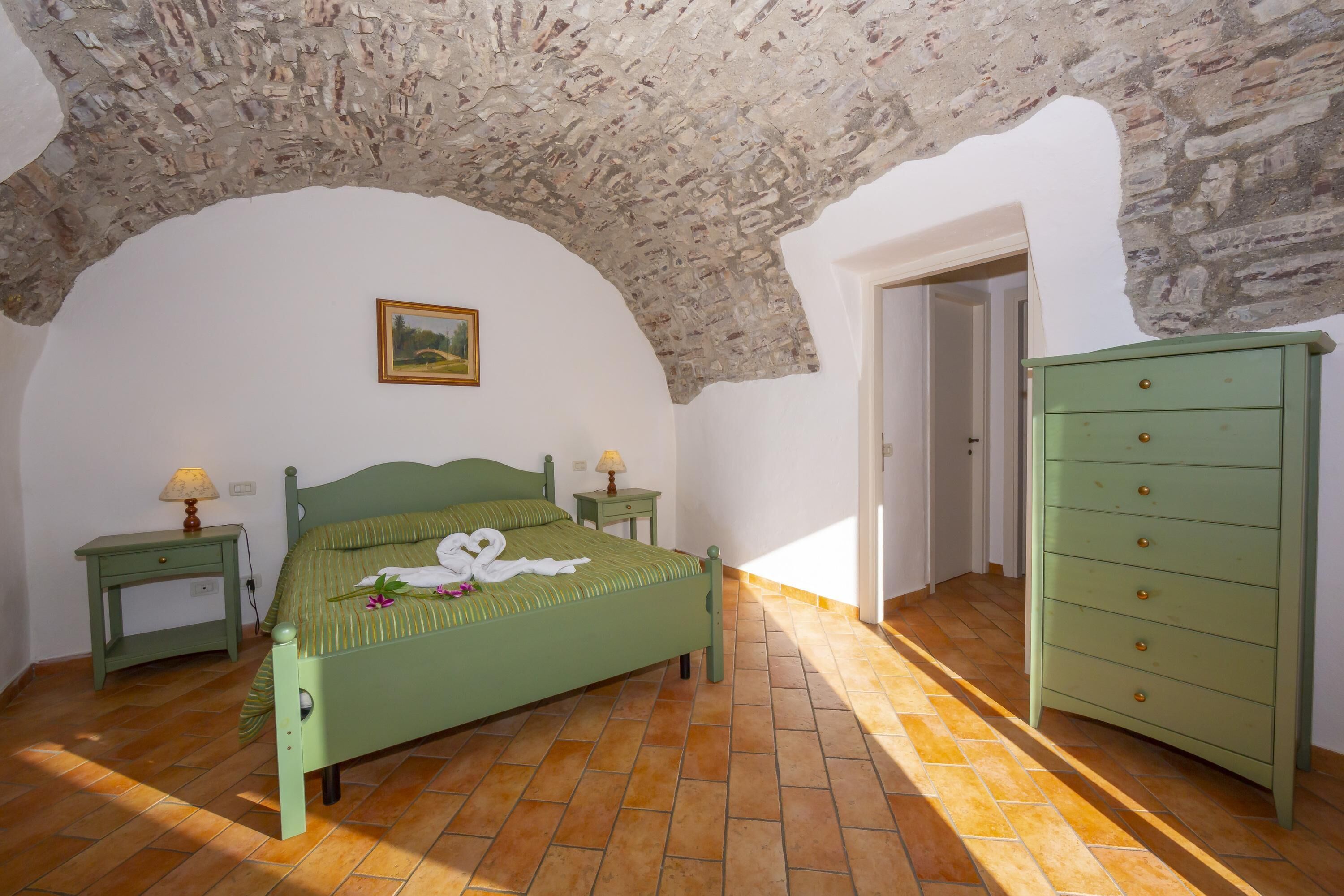 A bedroom with a stone ceiling, a green bed, a nightstand, a dresser, and a painting on the wall.