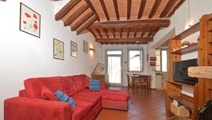 Un salon avec un canapé rouge, des poutres en bois au plafond, un téléviseur posé sur un meuble en bois, et un coin repas à l’arrière-plan.