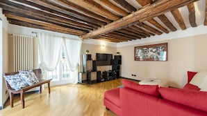 Un salon avec un plafond en bois, un canapé rouge, un banc en bois et un téléviseur posé sur un meuble noir.