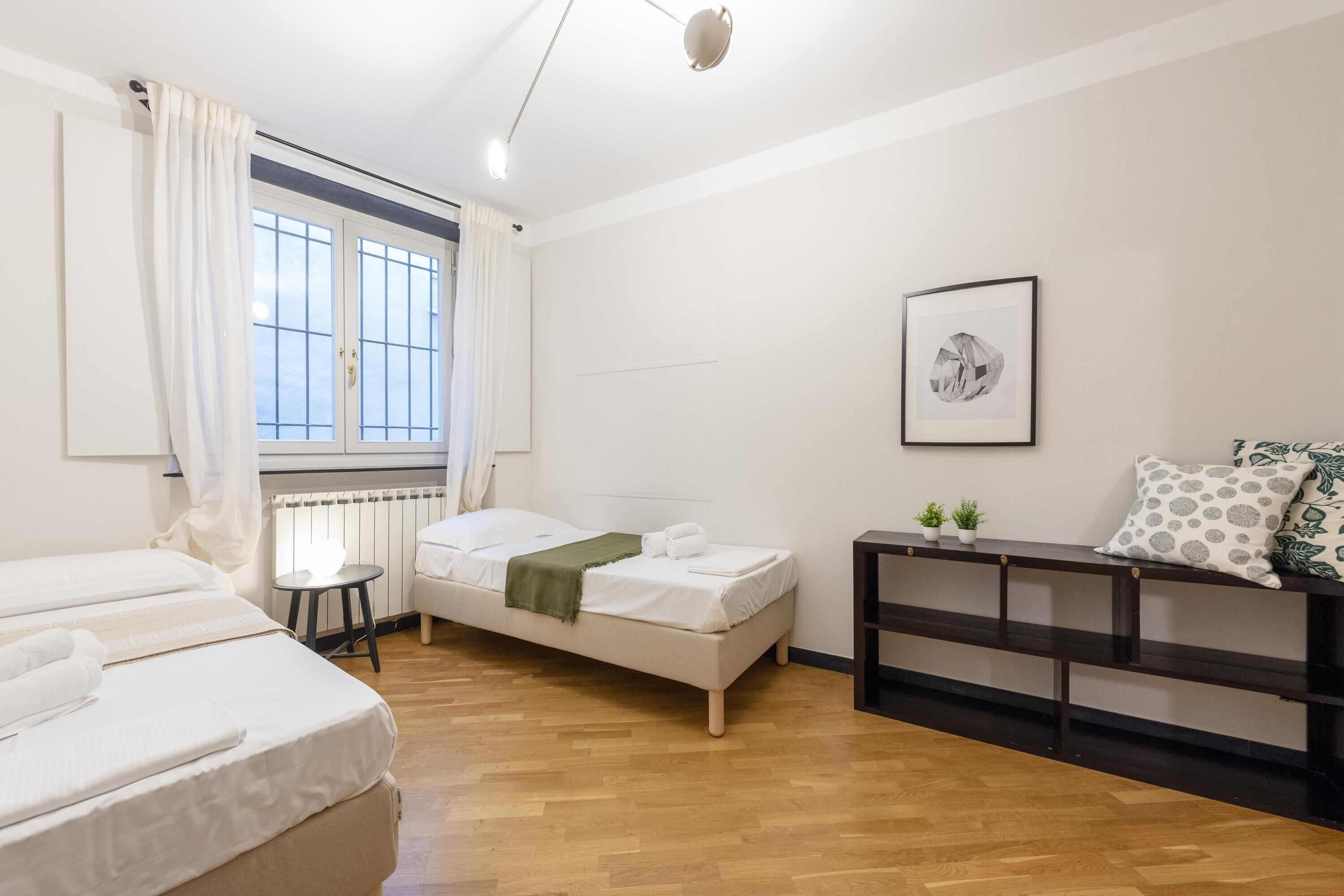 A hotel room with two beds, a small table, a bench, and a window with curtains.