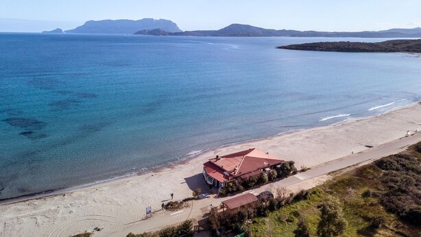 An aerial view of a coastal property with a beach, a red-roofed building, and distant mountains. - Le Dimore di Nettuno 1 With Pool (Olbia)