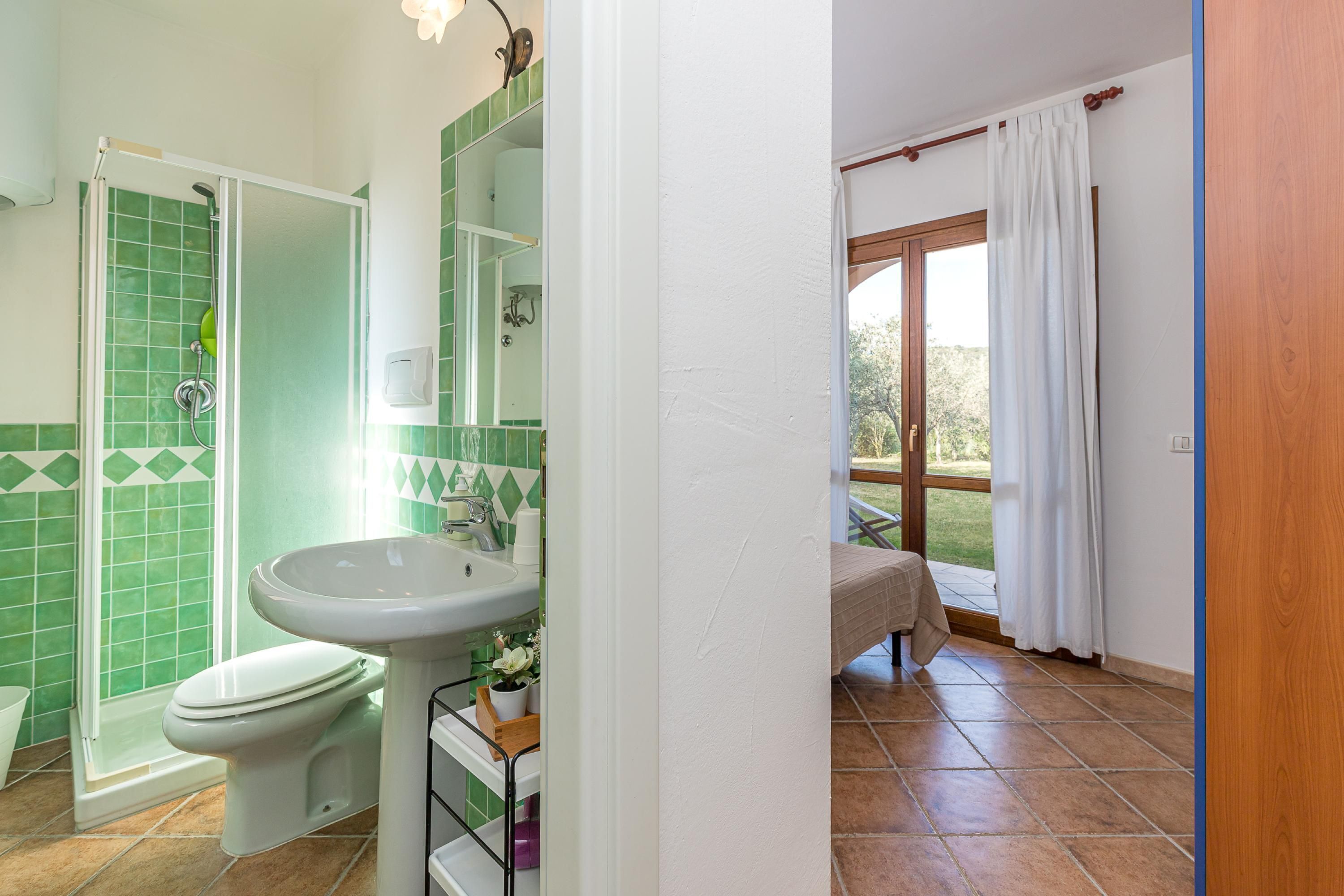 A bathroom with a shower, toilet, and sink, green tiles, and a view of the outdoors.