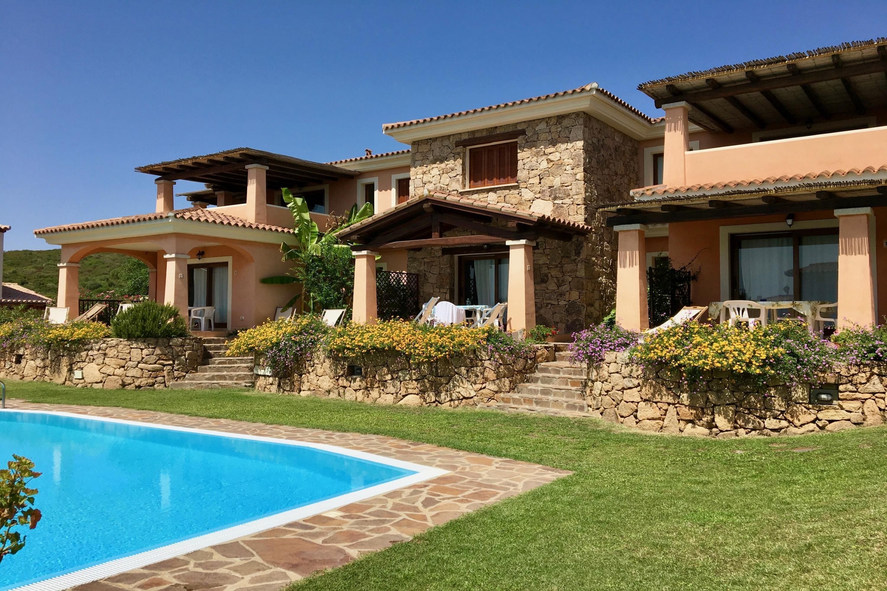 A poolside view of a resort with stone and stucco buildings, a swimming pool, and a well-maintained lawn.