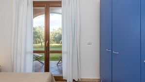 A bedroom with a bed, a blue wardrobe, and a sliding glass door leading to a patio with a lawn and a chair. - Le Dimore di Nettuno 1 With Pool (Olbia)