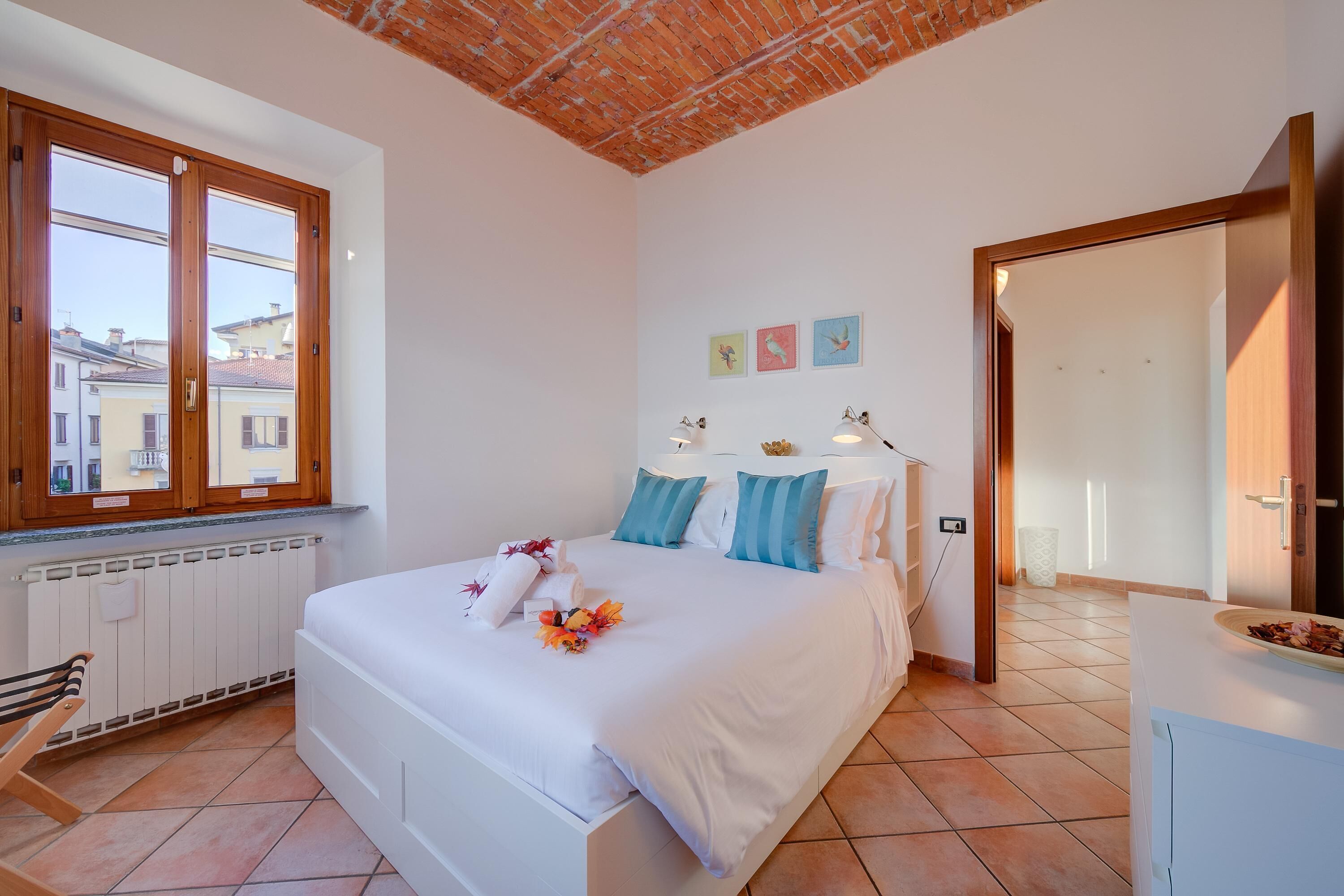 A bedroom with a large bed, a window with wooden frames, a radiator, and a door leading to another room.