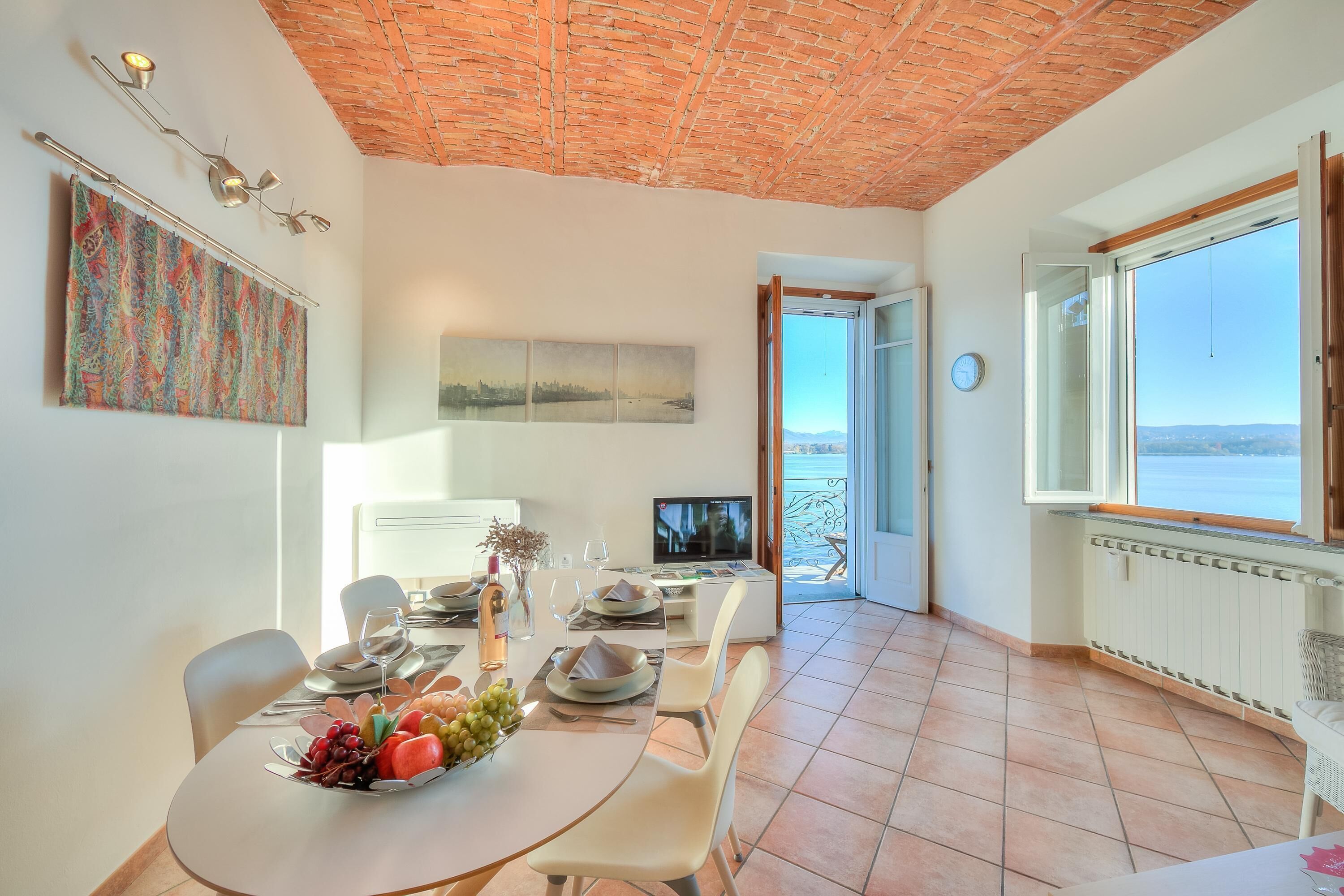 A dining area with a table set for four, a television, and a view of the sea through an open door.