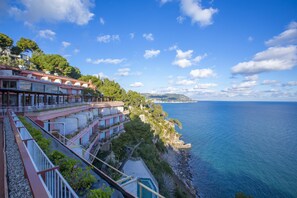 A coastal hotel with balconies overlooking the sea, a clear sky, and a distant view of a peninsula.