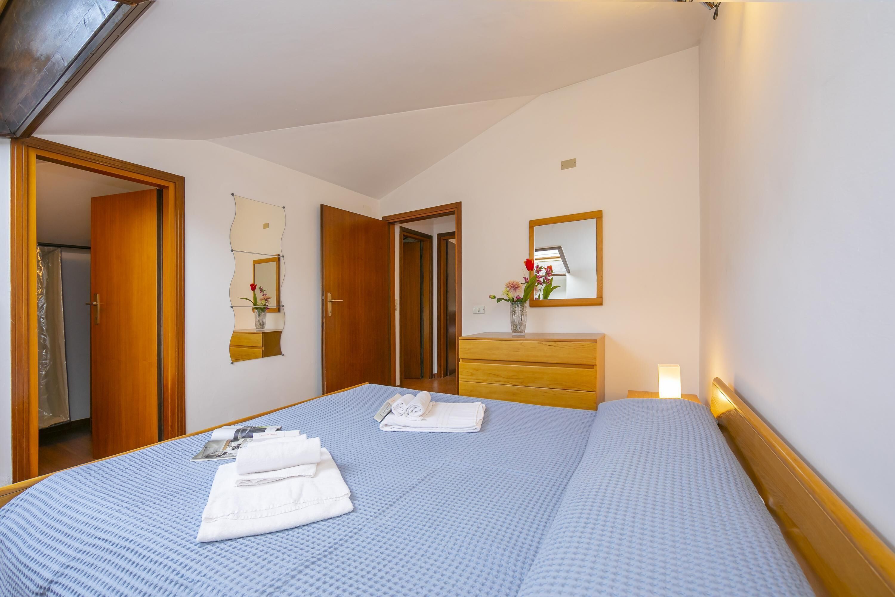 Une chambre d’hôtel avec un grand lit, une commode en bois, un miroir et un petit vase avec des fleurs.